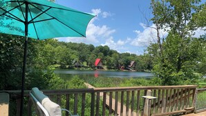 A wooden deck with a lounge chair and a table, overlooking a lake with houses in the background. - Lakefront Aframe Cabin in Hardy on Kiwanie Lake with kayaks and private dock (Hardy)