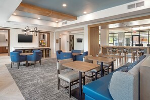 Un moderno vestíbulo de hotel con una zona de bar, diferentes disposiciones de asientos y una cocina visible al fondo.