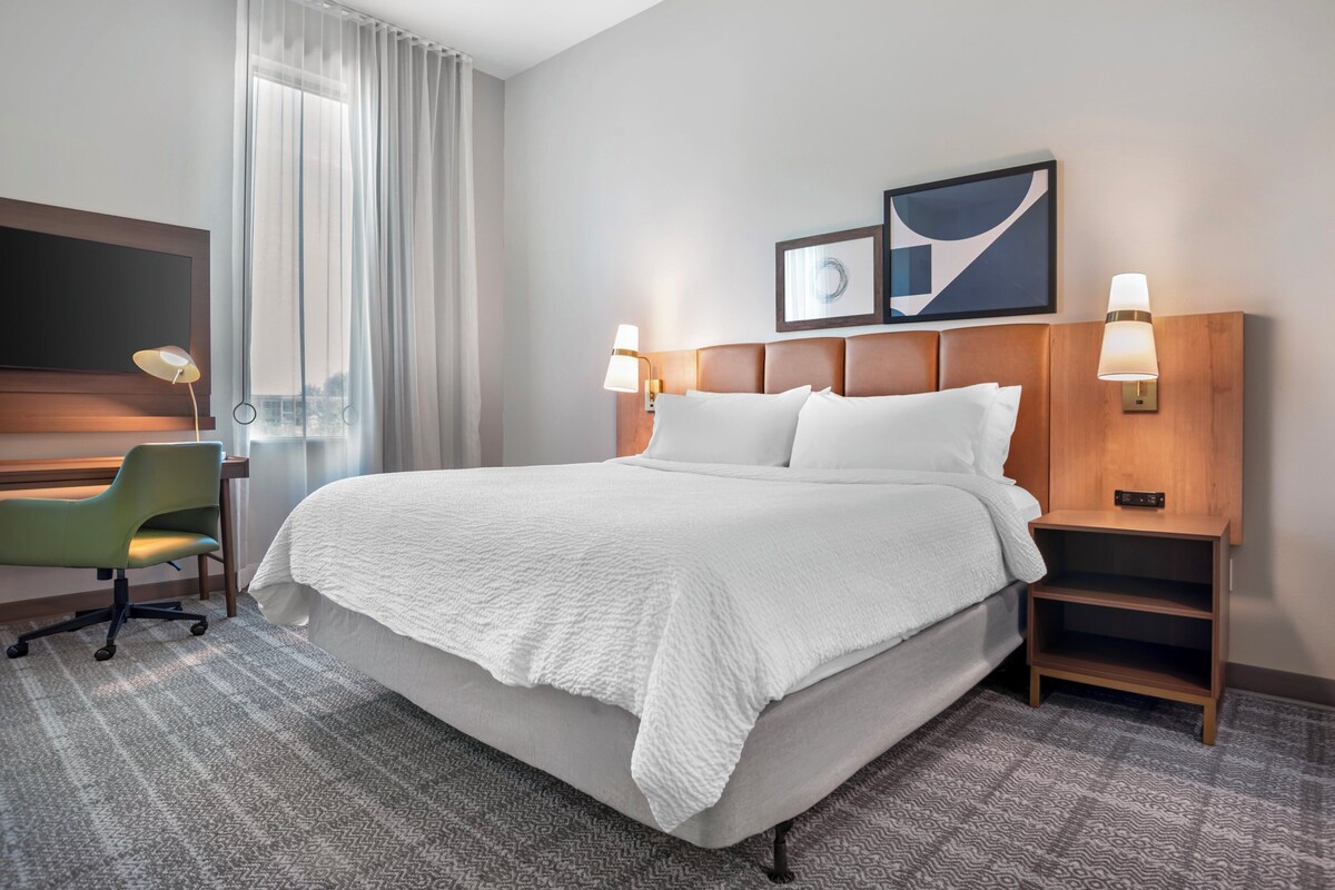 A hotel room with a bed, a desk, a chair, two bedside lamps, and artwork on the wall.