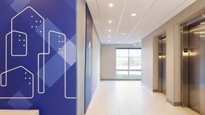 A modern hotel corridor with a blue wall featuring geometric shapes, a white bench, and an elevator. - Holiday Inn Express & Suites Charlotte Airport Area by IHG (Charlotte)