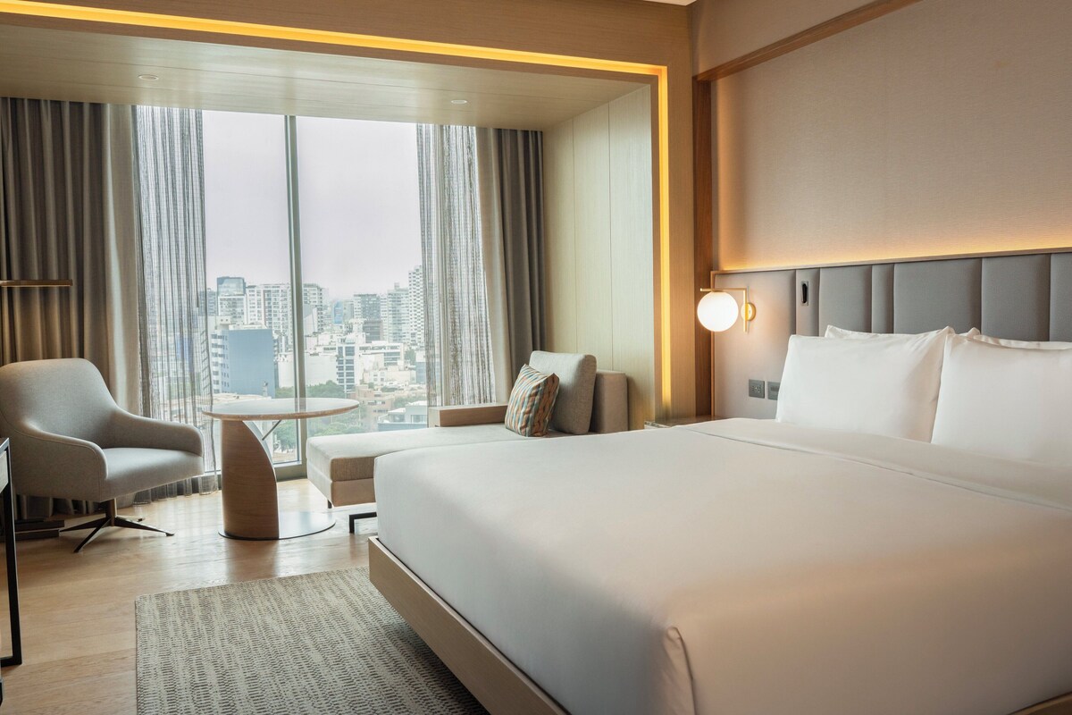 A modern hotel room with a large bed, a seating area with a table, and a view of the cityscape through large windows.
