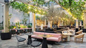 An outdoor seating area with cushioned benches and tables, surrounded by greenery and string lights.