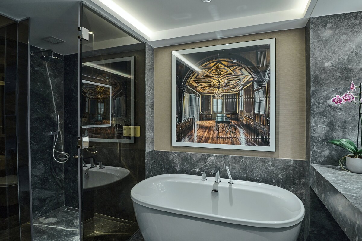 A modern bathroom with a freestanding bathtub, a large mirror, and a framed picture of a grand interior.
