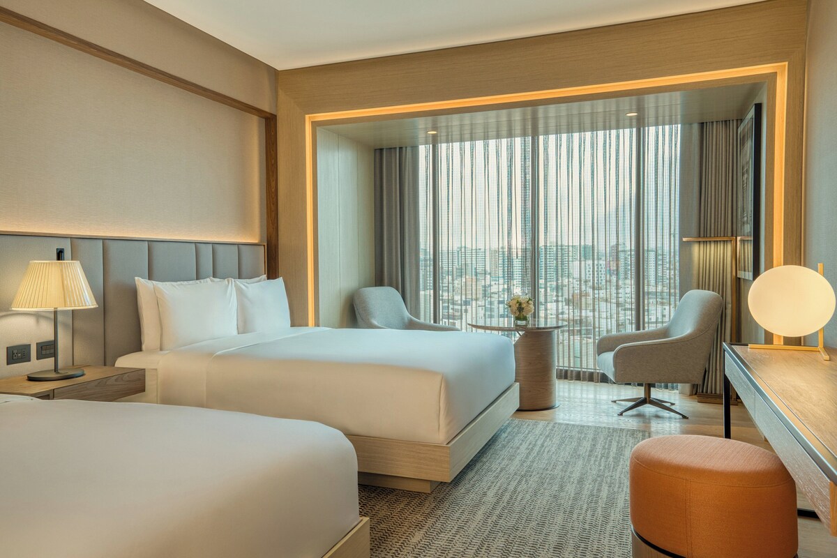 Une chambre d’hôtel avec deux lits, un bureau, une chaise et une vue sur la ville grâce à de grandes fenêtres.