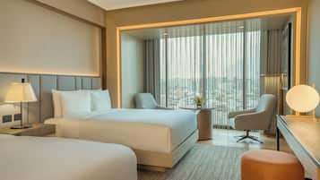 A hotel room with two beds, a desk, a chair, and a view of the city through large windows.