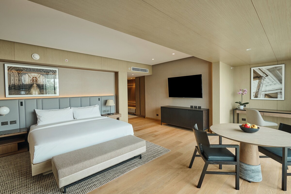 A modern hotel room with a large bed, a dining area with a round table, and a flat-screen TV mounted on the wall.