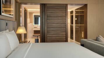 Una habitación de hotel con una cama grande, una mesita de noche con lámpara, un sofá y una puerta corredera que da al baño.