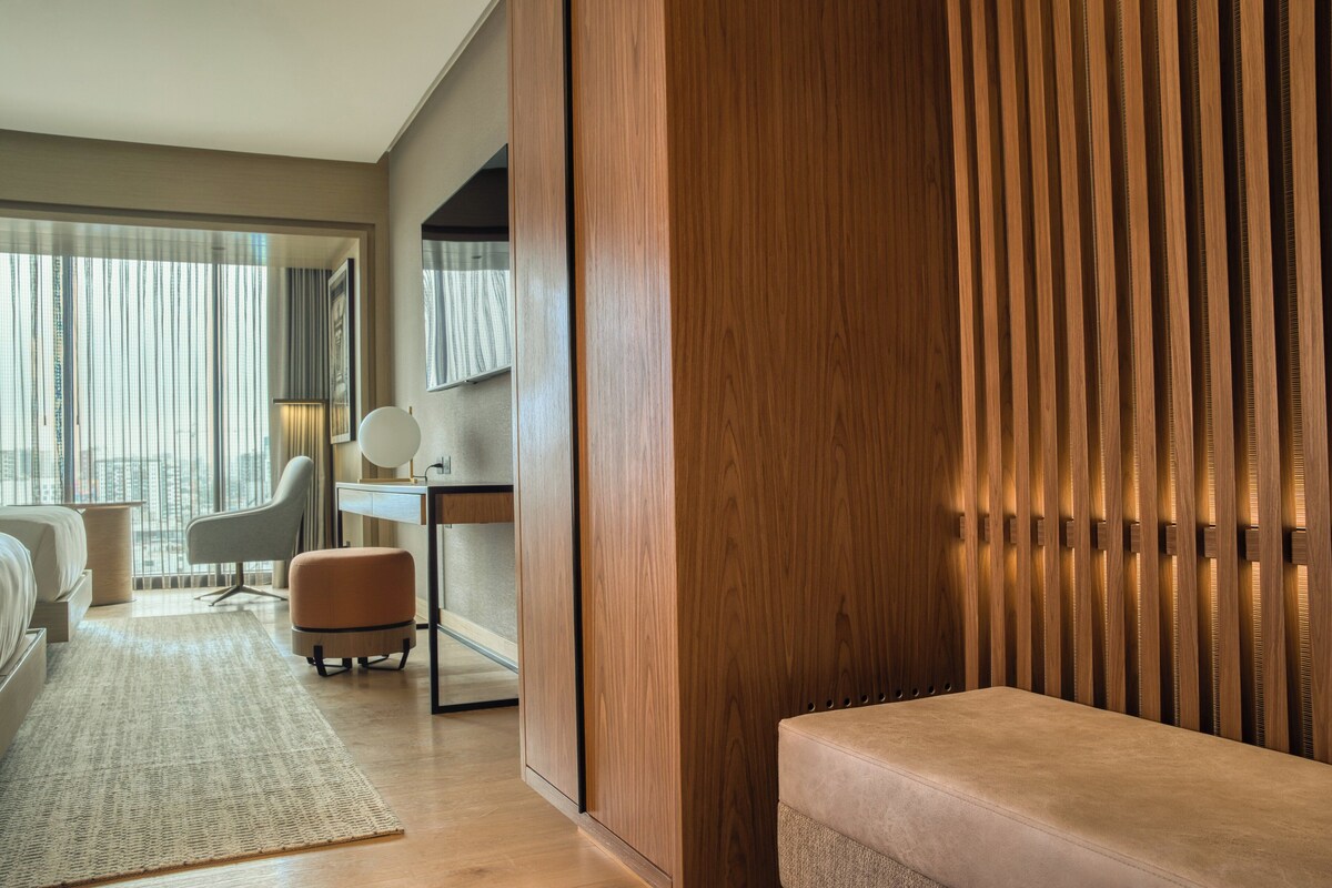 A modern hotel room with a wooden accent wall, a desk with a lamp, a sofa, and a view of the city through large windows.
