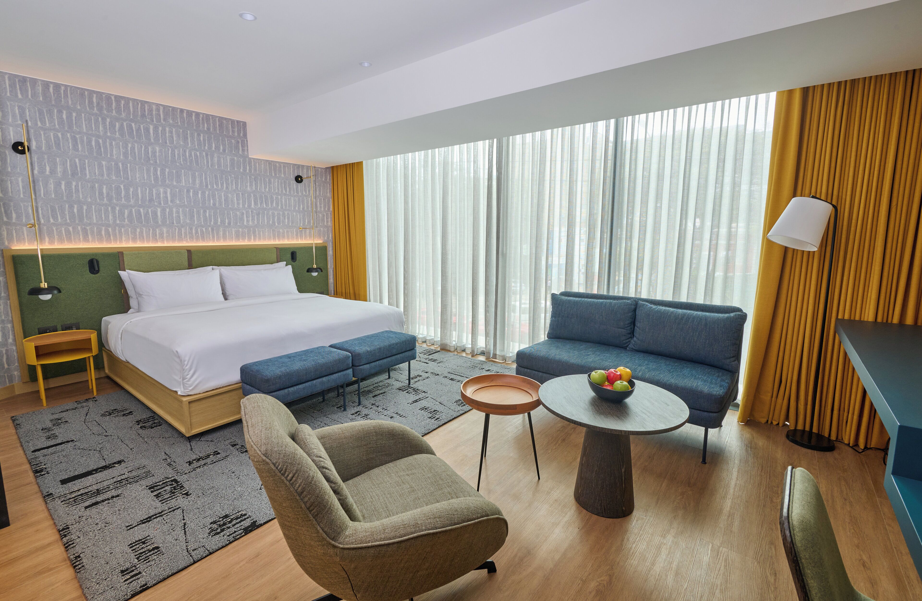Una habitación de hotel moderna con una cama, dos sofás, una mesita con fruta y un gran ventanal con cortinas.