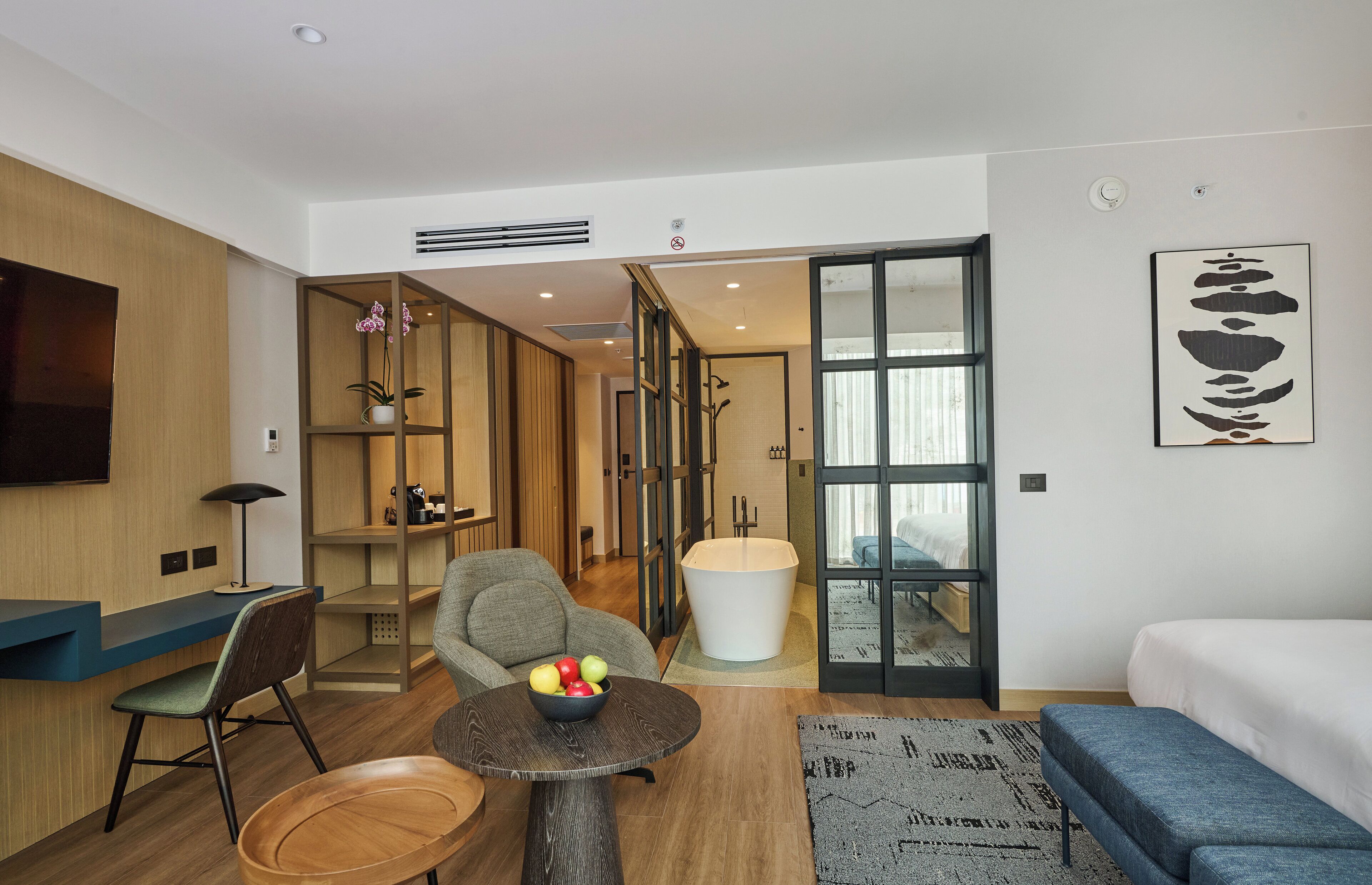 A modern hotel room with a flat-screen TV, a round wooden table with a bowl of fruit, a grey armchair, a blue sofa, and a large window.