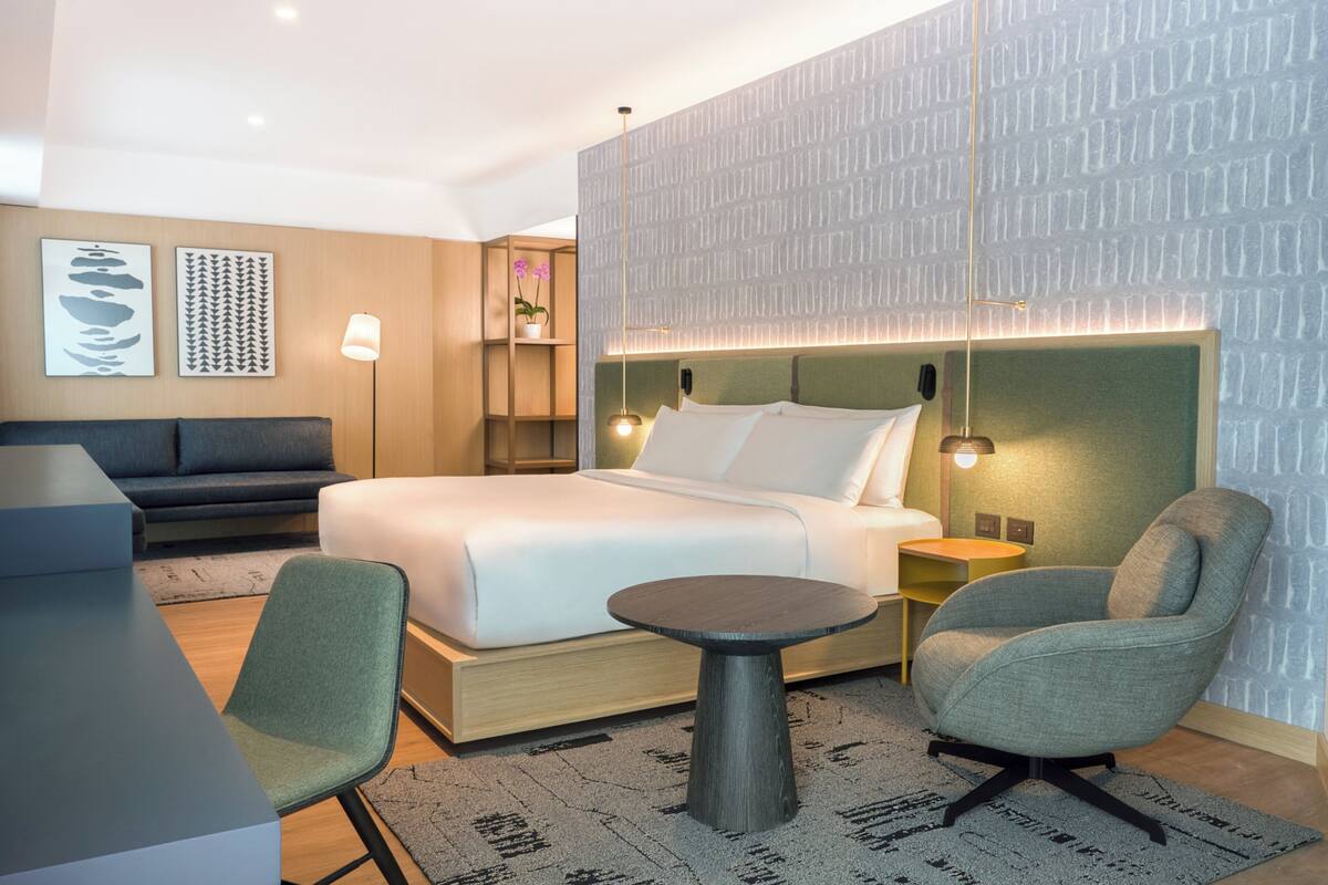 A modern hotel room with a large bed, a desk, two chairs, a small table, and a patterned wall.