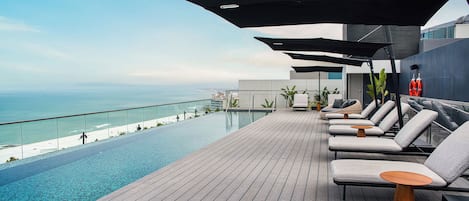 A rooftop infinity pool with lounge chairs and umbrellas, overlooking a beach.