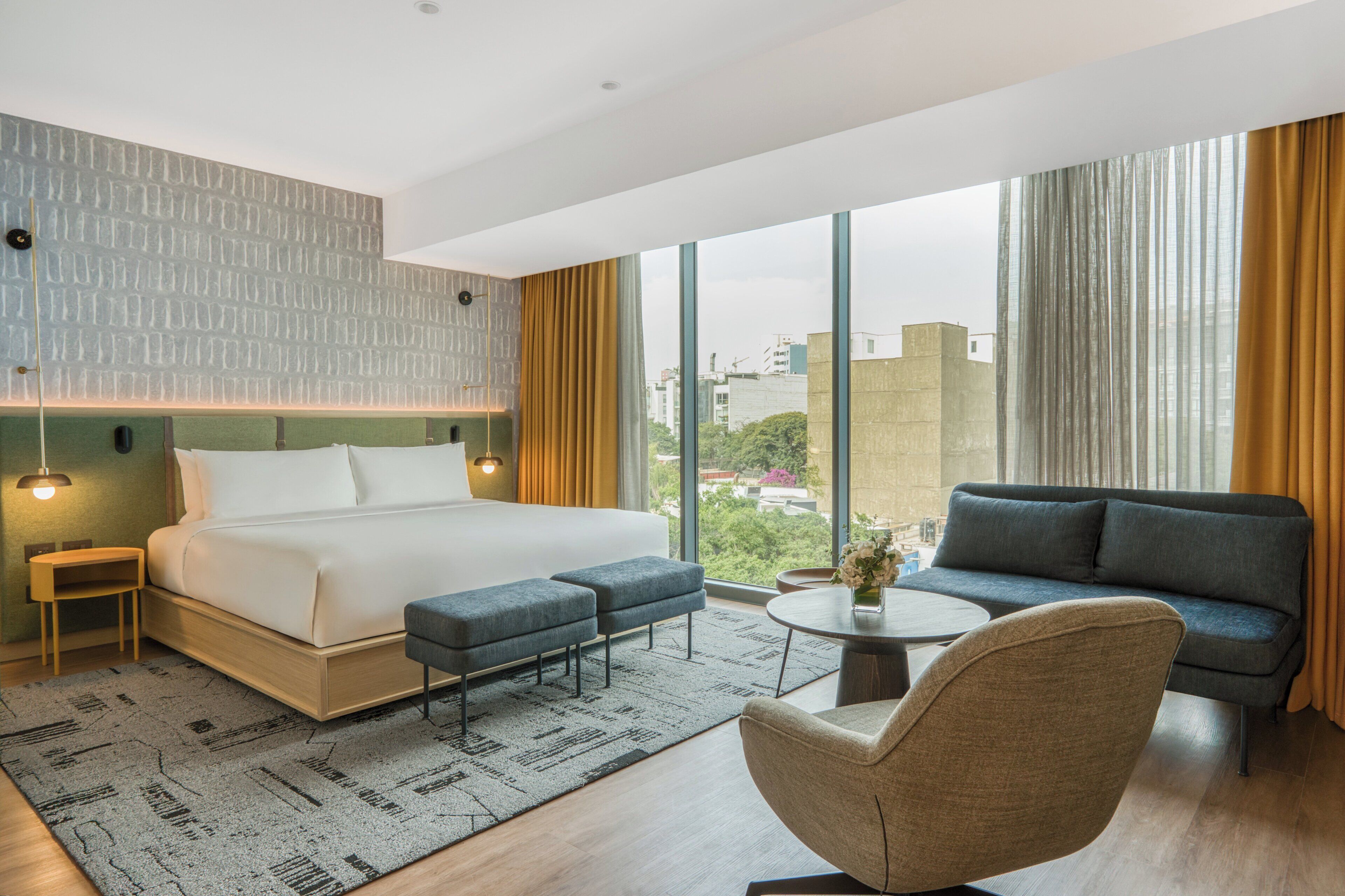 A modern hotel room with a large bed, a sofa, a small table, and a view of the city.