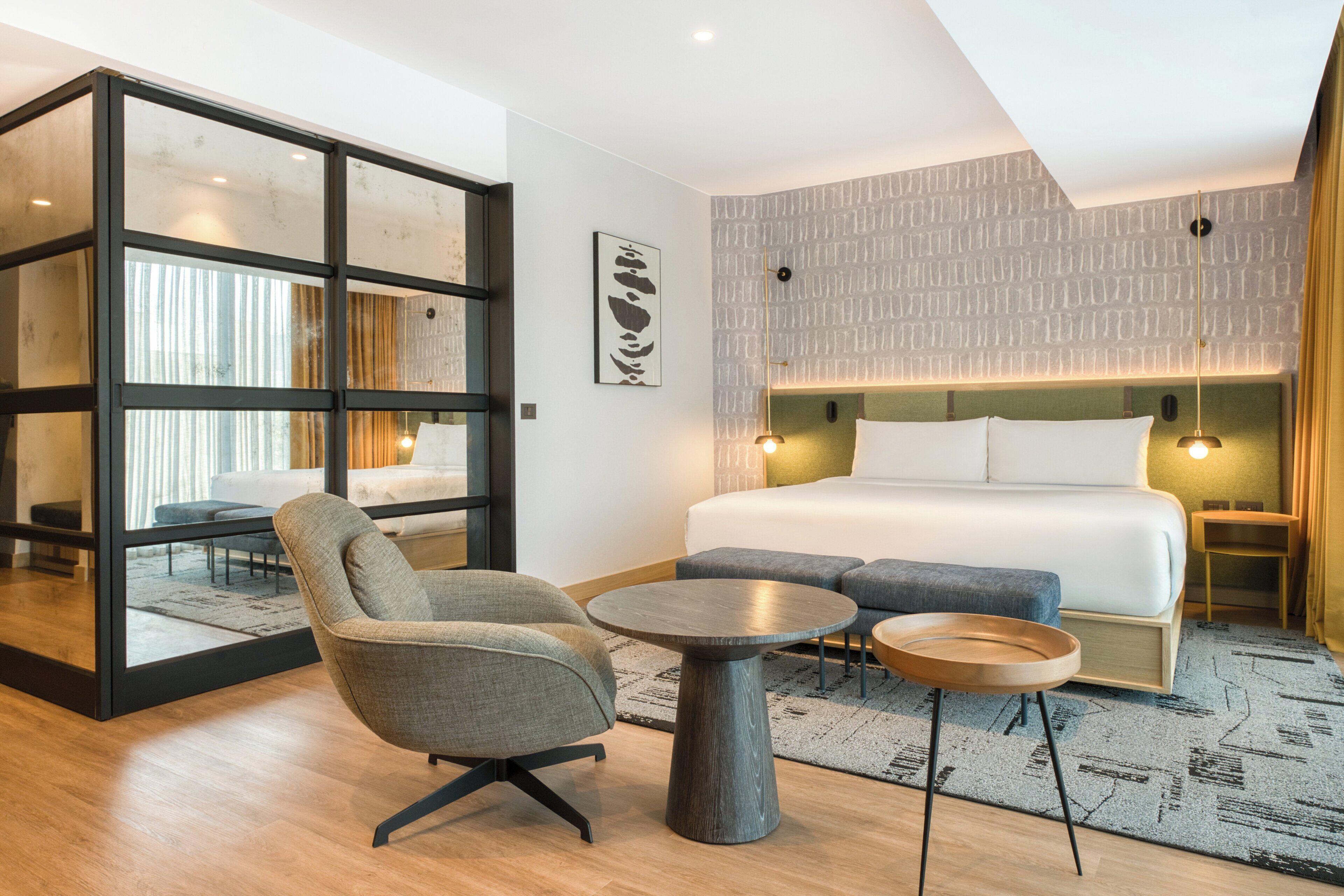 A modern hotel room with a large bed, a seating area with a chair and a round table, and a glass partition.