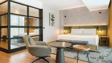 A modern hotel room with a large bed, a seating area with a chair and a round table, and a glass partition.
