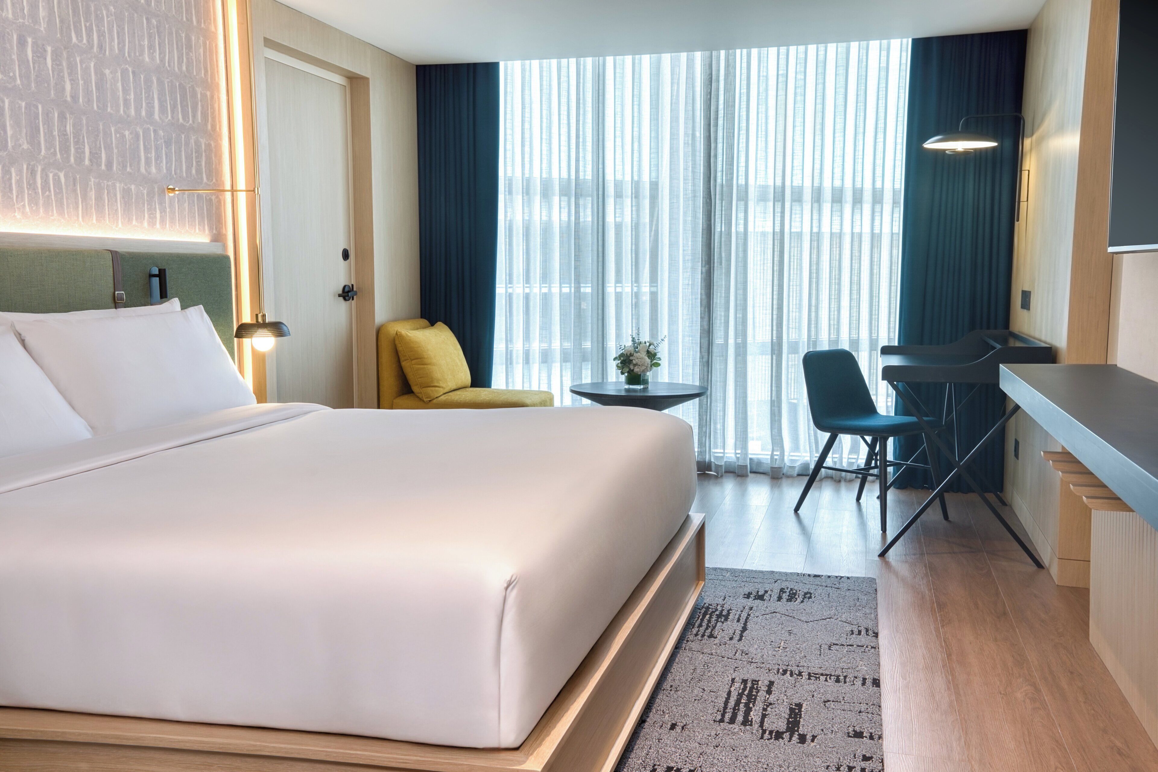 A hotel room with a large bed, a desk with two chairs, a small table with a vase, and a view of the city through sheer curtains.