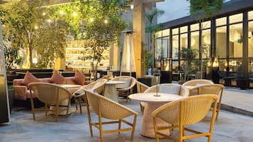 Un espacio al aire libre con sillas y mesas de mimbre, rodeado de plantas en macetas y una zona de bar con estantes llenos de botellas.