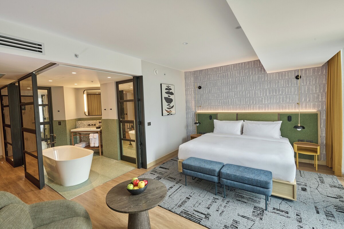 Habitación de hotel moderna con una cama grande, una bañera, una mesita con fruta y vistas al baño.