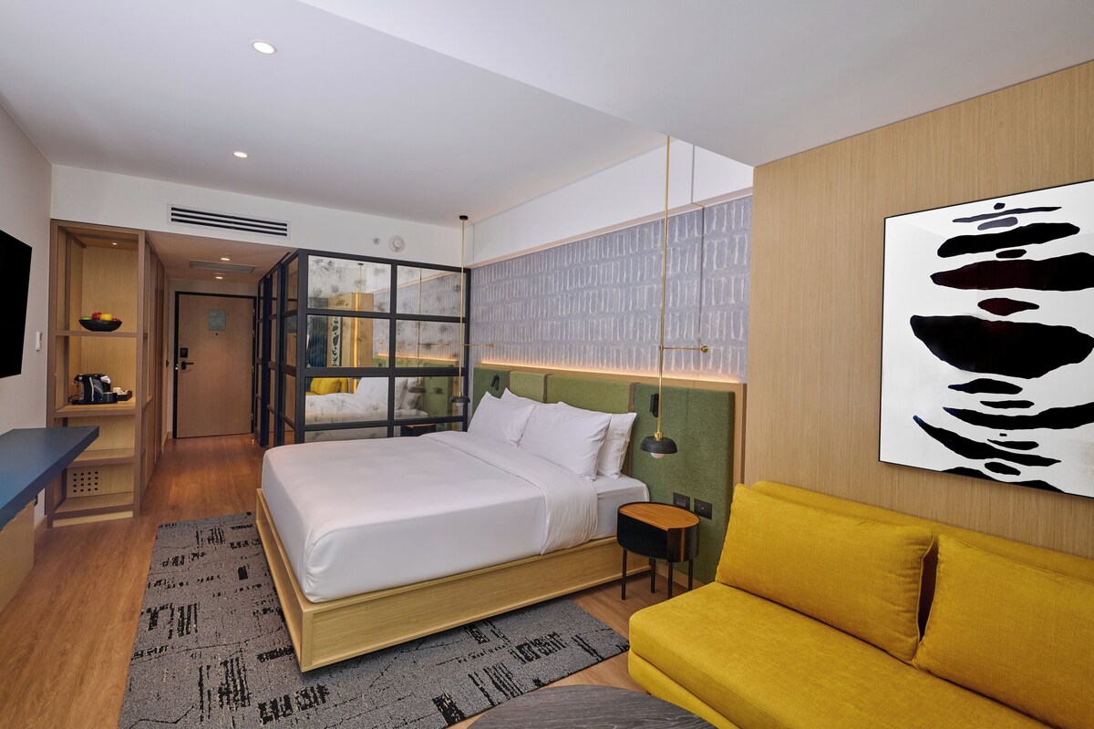 Una habitación de hotel moderna con una cama grande, un sofá amarillo, un televisor de pantalla plana y una original obra de arte en la pared.