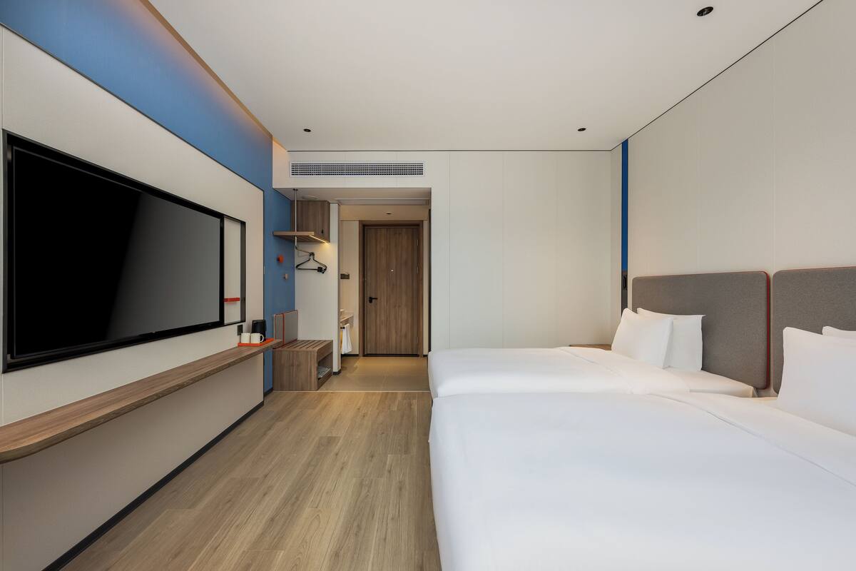 Une chambre d’hôtel moderne dotée d’une grande télévision à écran plat, d’une table de chevet en bois, d’un lit avec du linge de maison blanc et d’un dressing.