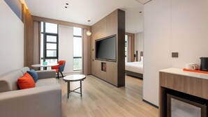 Een moderne hotelkamer met een bank, een klein rond tafeltje, een aan de muur bevestigde televisie en een kitchenette met een koffieapparaat.
