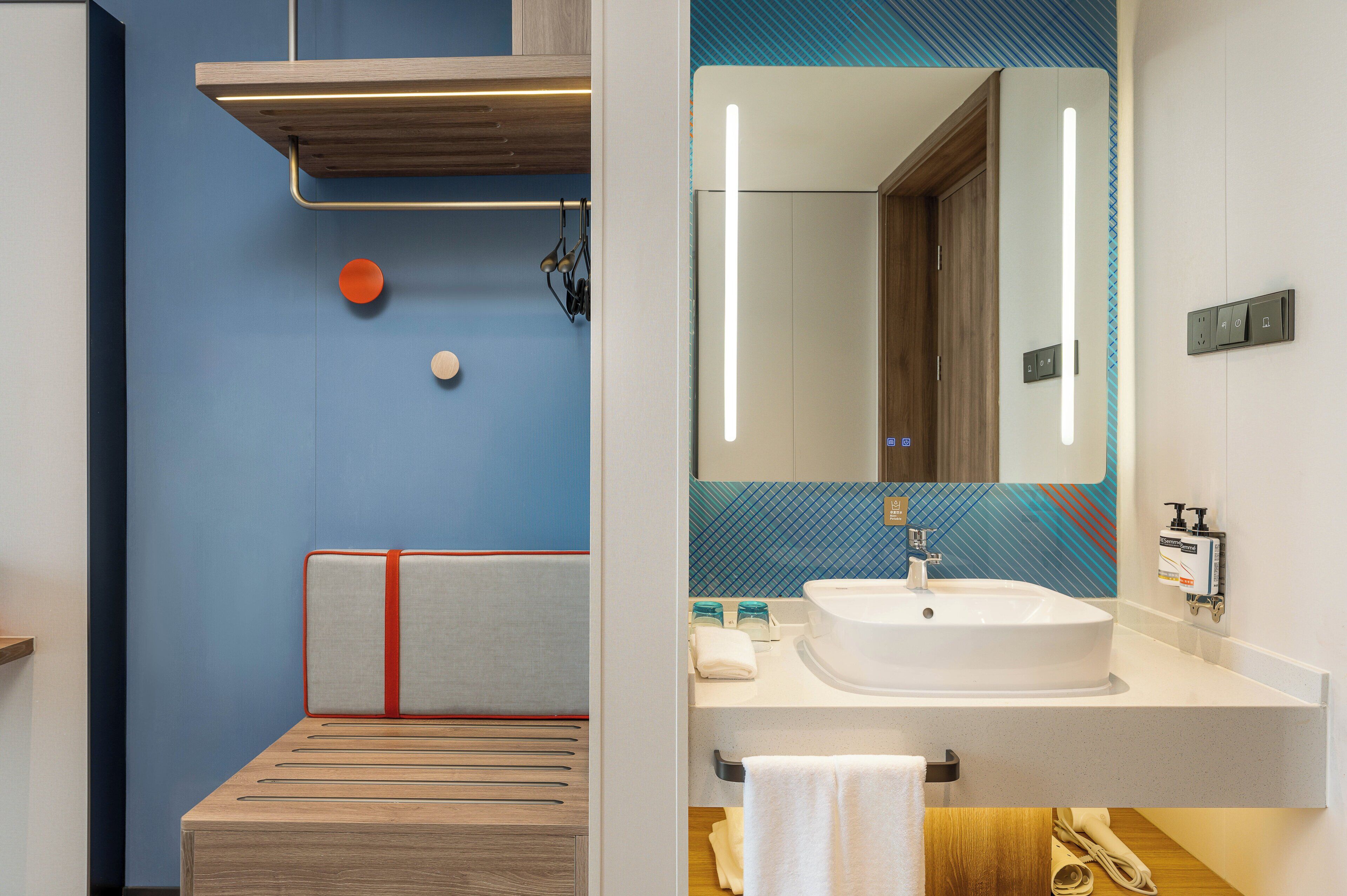 Une salle de bain moderne avec un mur d’accent bleu, un banc en bois, un lavabo blanc et un miroir avec un éclairage vertical.