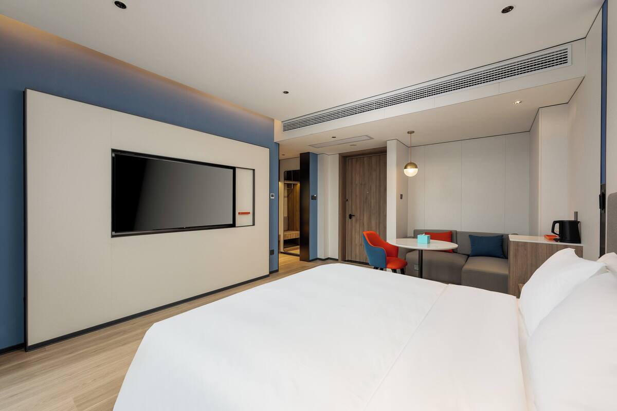 Une chambre d’hôtel moderne dotée d’un grand lit, d’un téléviseur à écran plat fixé au mur, d’une petite table avec une chaise et d’un canapé agrémenté de coussins.