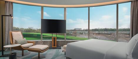 A modern hotel room with a large bed, a seating area with a chair and ottoman, a flat-screen TV, and a view of a golf course.