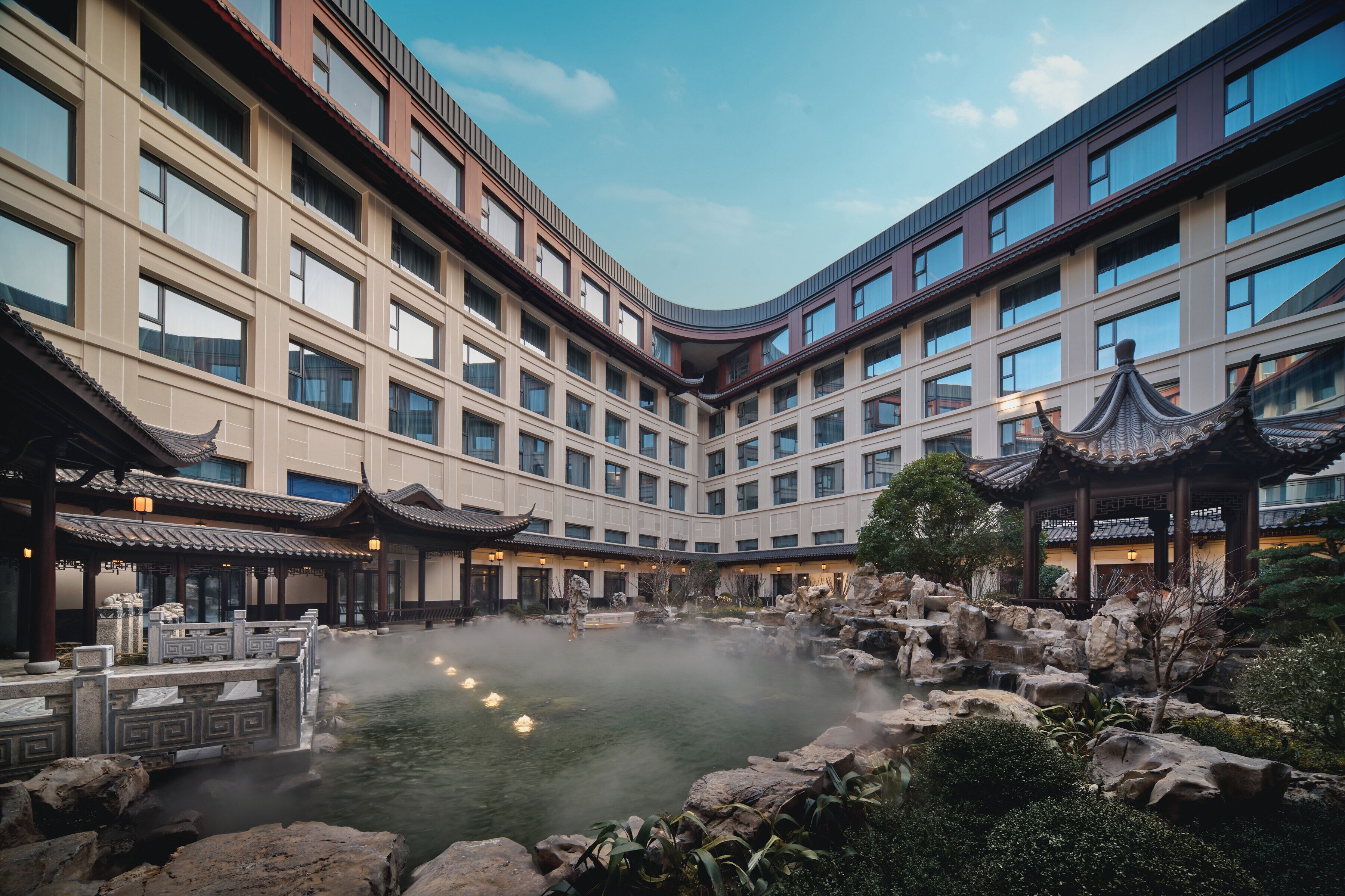 A hotel courtyard with a traditional pavilion, a hot spring, and a modern building with multiple windows.