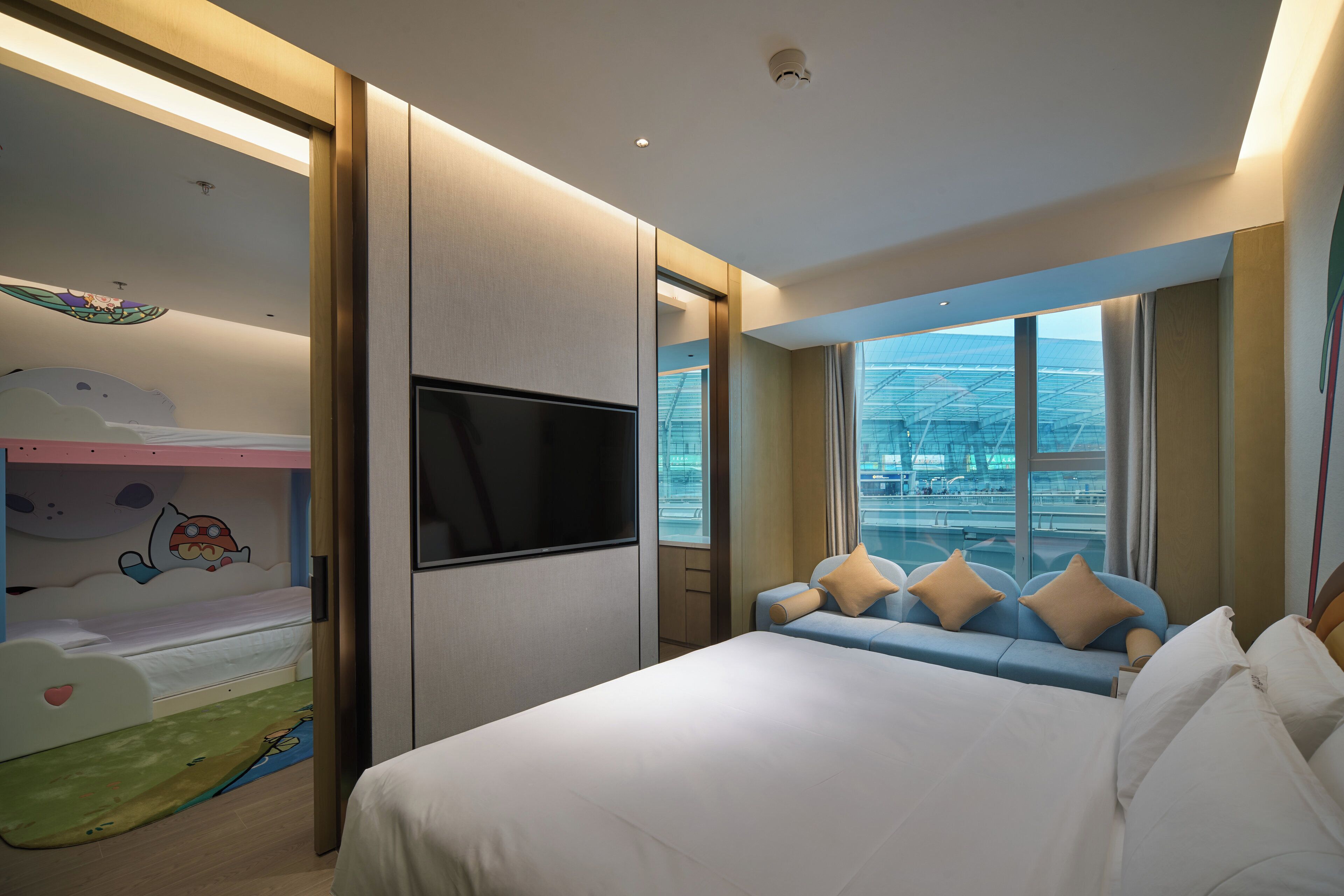 Une chambre d’hôtel comprenant un lit, une télévision, un canapé avec des coussins, un lit superposé pour enfants et une vue sur le paysage urbain grâce à de grandes fenêtres.