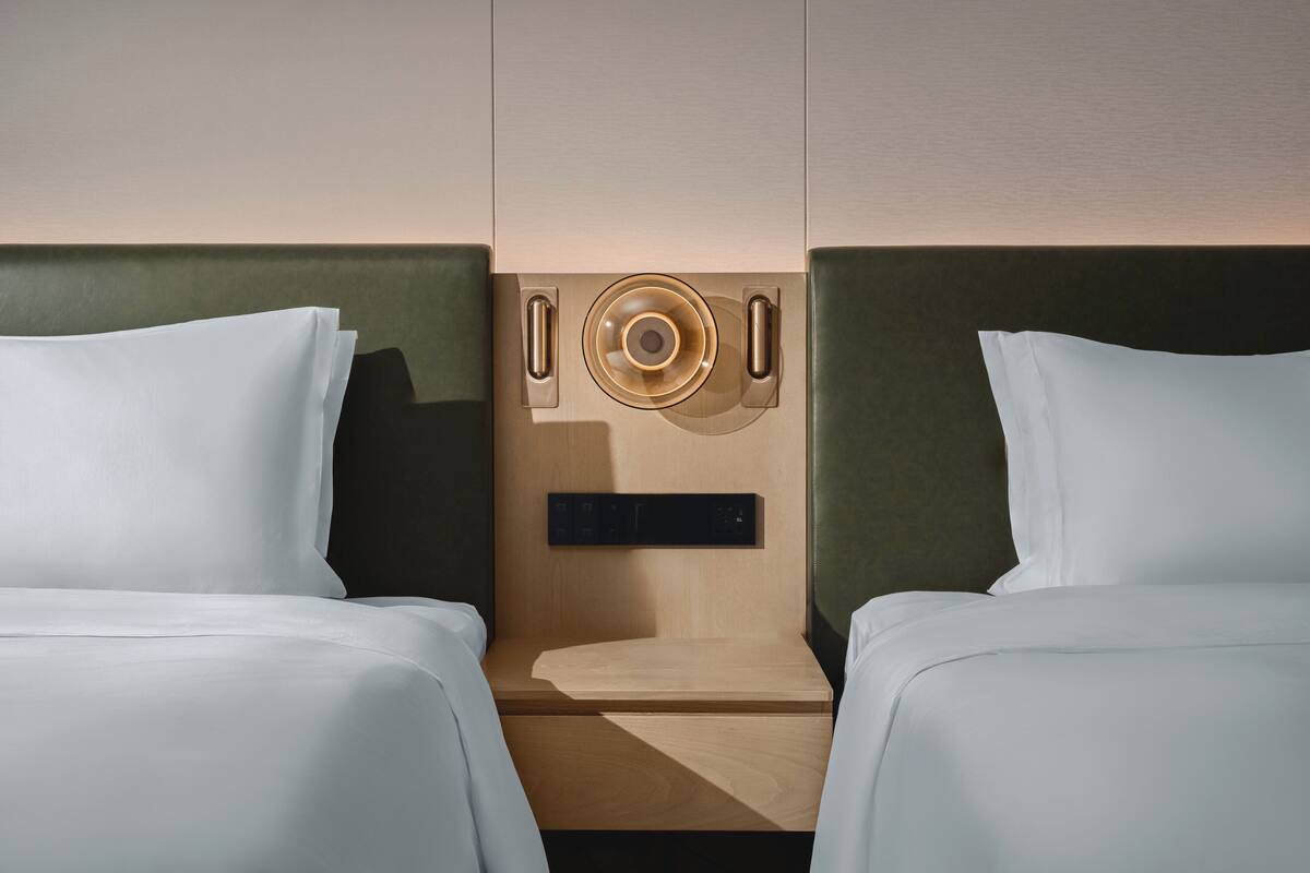 Habitación de hotel con dos camas, cabecero de madera, una mesita pequeña y un panel de control en la pared.