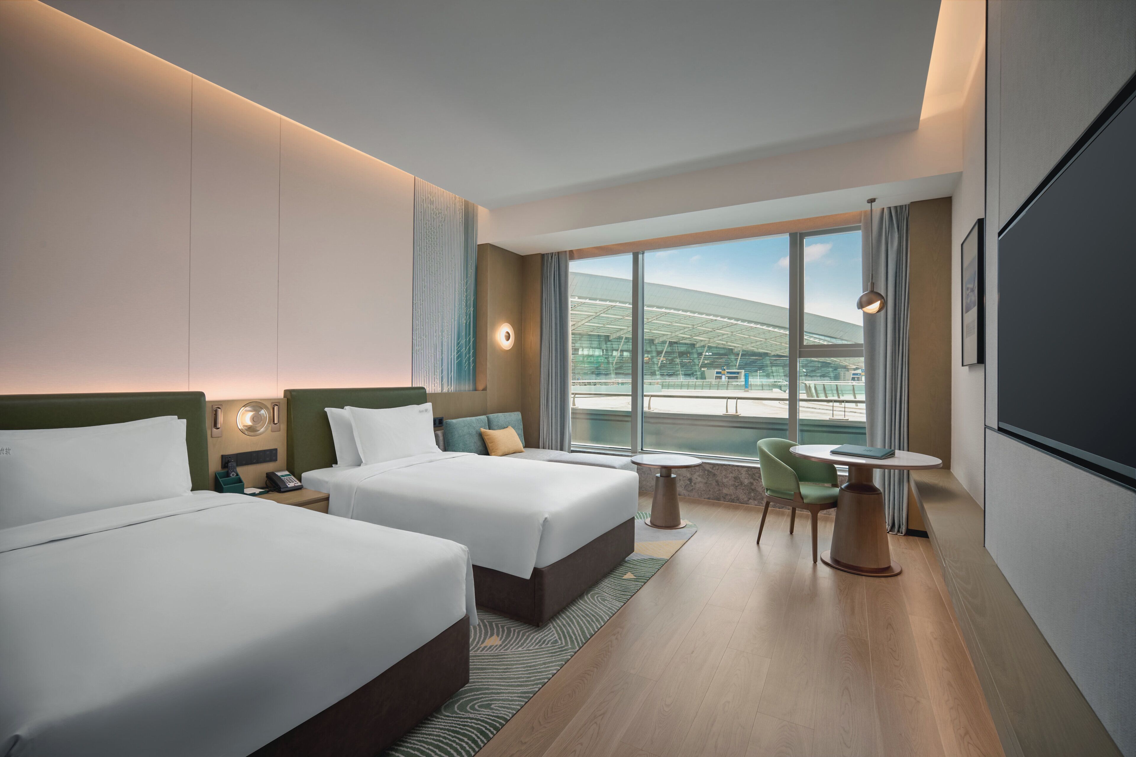 Habitación de hotel con dos camas, una mesita, una silla y un amplio ventanal con vistas a un edificio moderno.