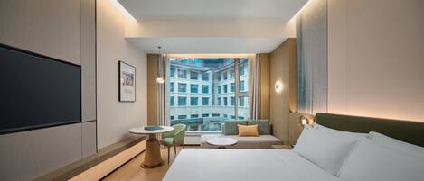 A modern hotel room with a large bed, a small table, a sofa, and a TV on the wall.