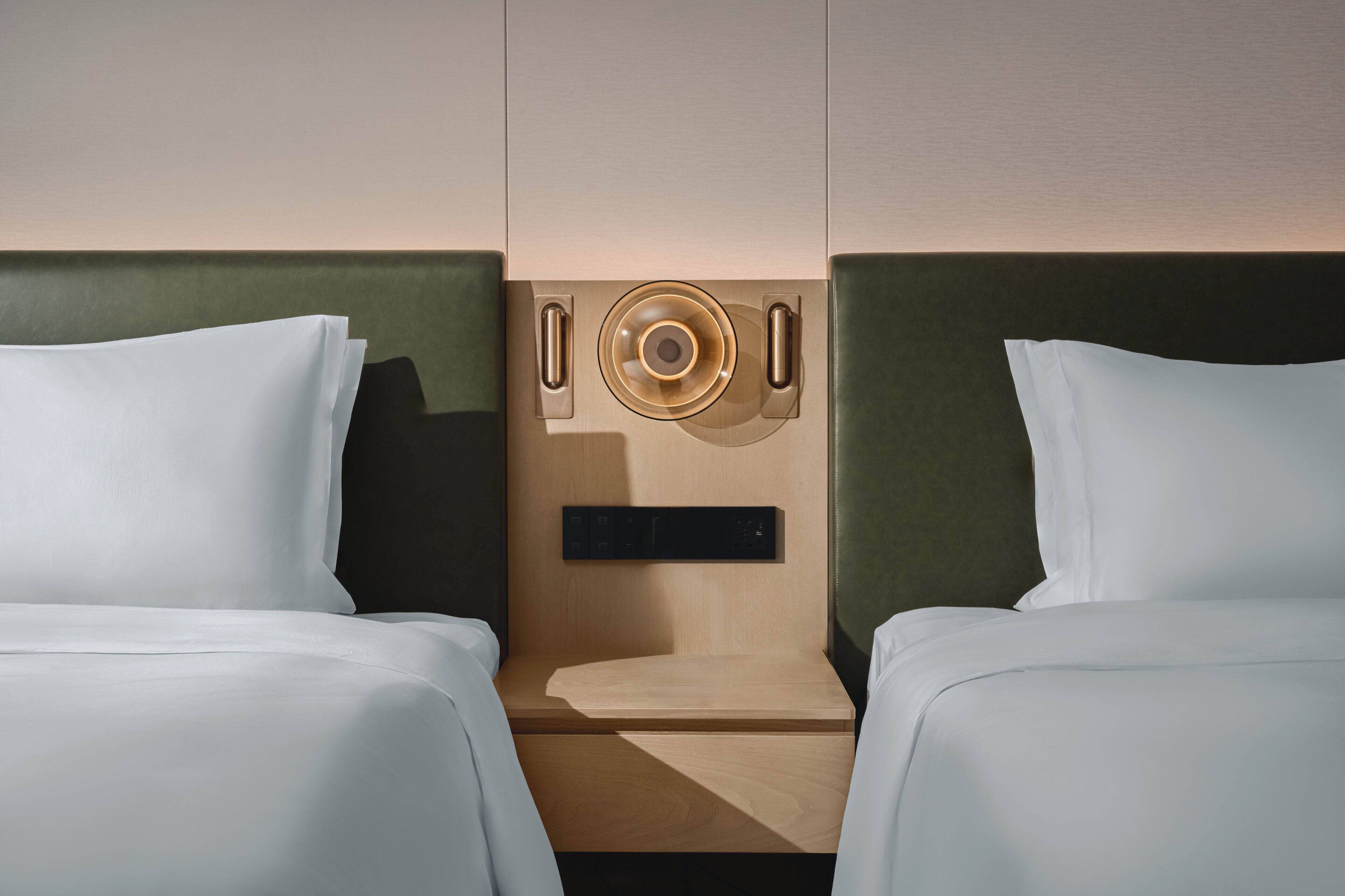 Habitación de hotel con dos camas, cabecero de madera, una mesita pequeña y un panel de control en la pared.