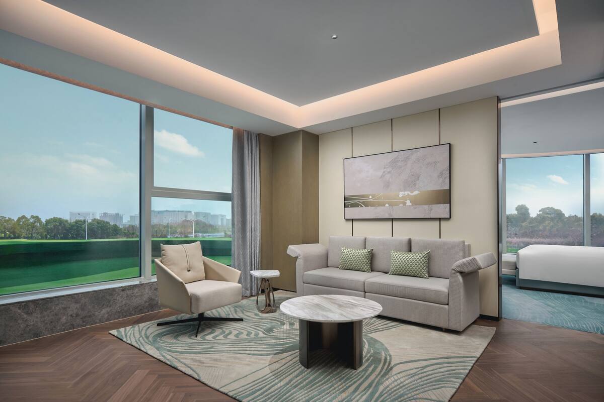 A modern hotel room with a large window, a sofa, a coffee table, and a painting on the wall.