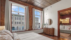 Une chambre à coucher avec un grand lit, deux fenêtres avec des rideaux, une commode en bois et une vue sur un bâtiment à l’extérieur.