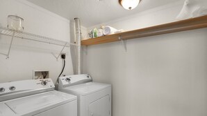 Une buanderie équipée de deux machines à laver, d’un étendoir et d’une étagère où sont rangées des serviettes et des bouteilles.