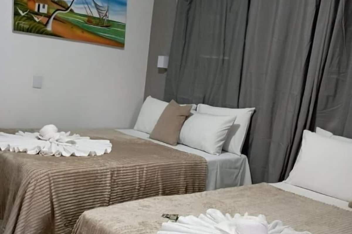 Hotelkamer met twee bedden, elk voorzien van een beige sprei en witte kussens. Aan de muur hangt een schilderij van een tropisch landschap, en erboven bevinden zich airconditioners.