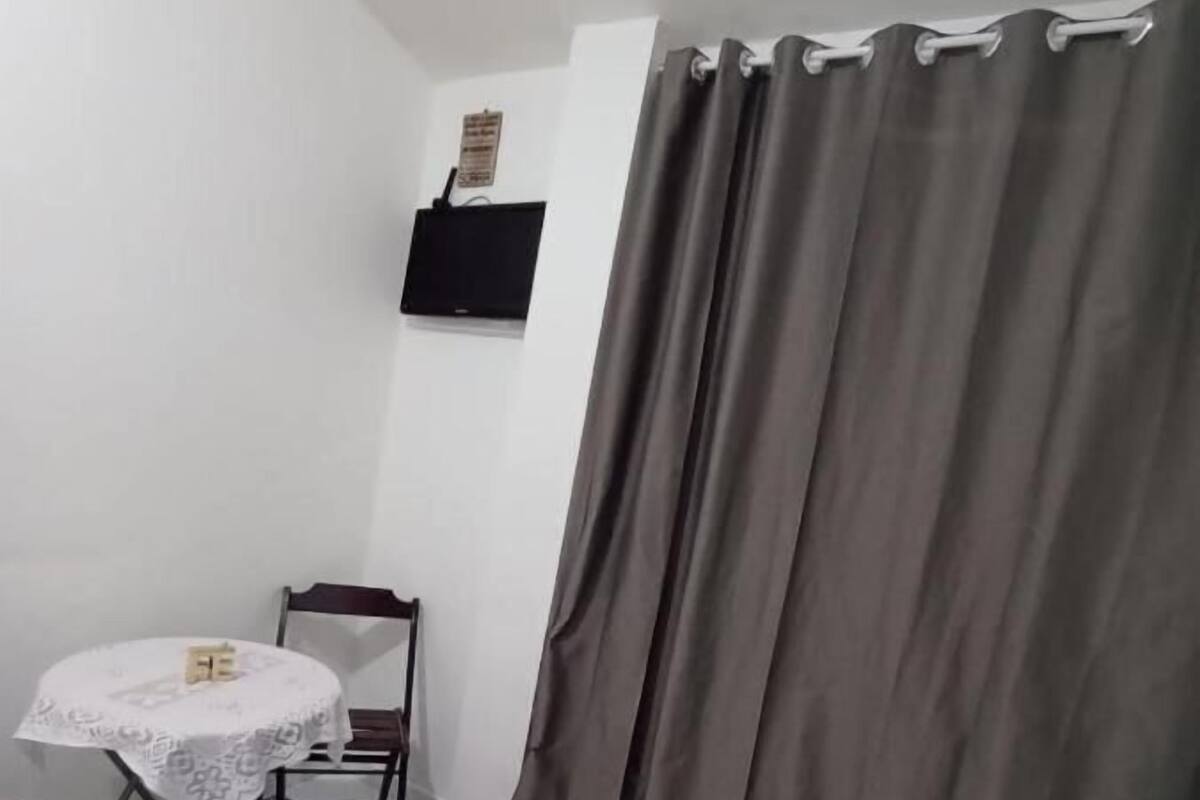 Een kleine kamer met een vloer van witte tegels, een ronde tafel met een witte tafelkleed, een grijze gordijn en een klein, groen vloermatje.