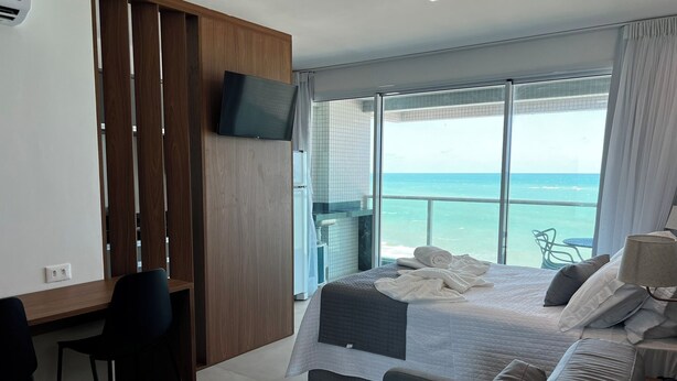 A modern hotel room with a large bed, a desk, a TV, and a balcony with a view of the ocean. - RN STUDIO - APTO 1008 (Maceio)