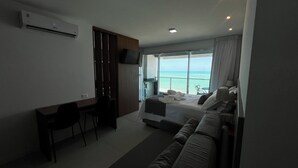 A hotel room with a bed, a desk, a chair, a TV, and a large window with a view of the ocean. - RN STUDIO - APTO 1008 (Maceio)