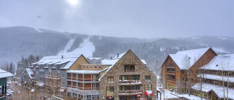A snow-covered resort with multiple buildings, including a stone building with a balcony and a wooden building with a ski slope in the background.