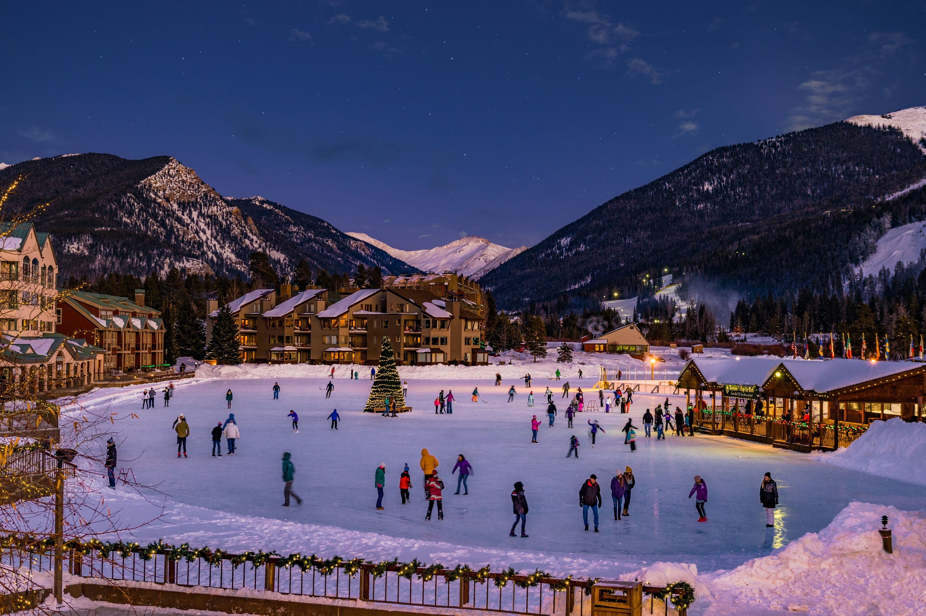 A snow-covered outdoor ice skating area with buildings and mountains in the background.