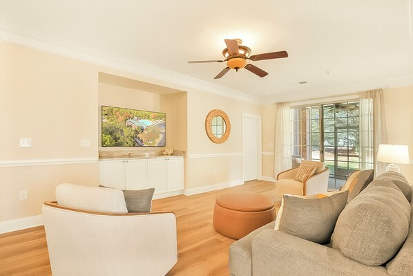 A spacious living room with a ceiling fan, a large window with curtains, a television mounted on the wall, and a view of the outdoors.