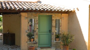 A small, single-story building with a green door, stone walls, and a tiled roof. There are two potted plants on the steps and a small cabinet to the left. - Vacation apartment with large pool in the hills of the bay of St. Tropez (La Garde-Freinet)