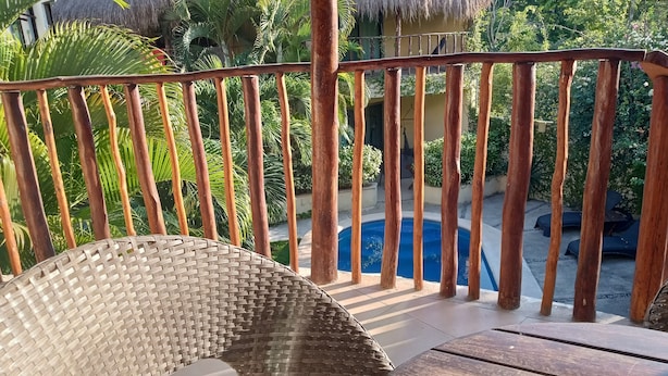 A wooden deck with a wicker chair, a pool, and a thatched-roof structure in the background. - Wonderful space with 2 beds, overlooking the pool, downtown Tulum- Huanal H (Tulum)