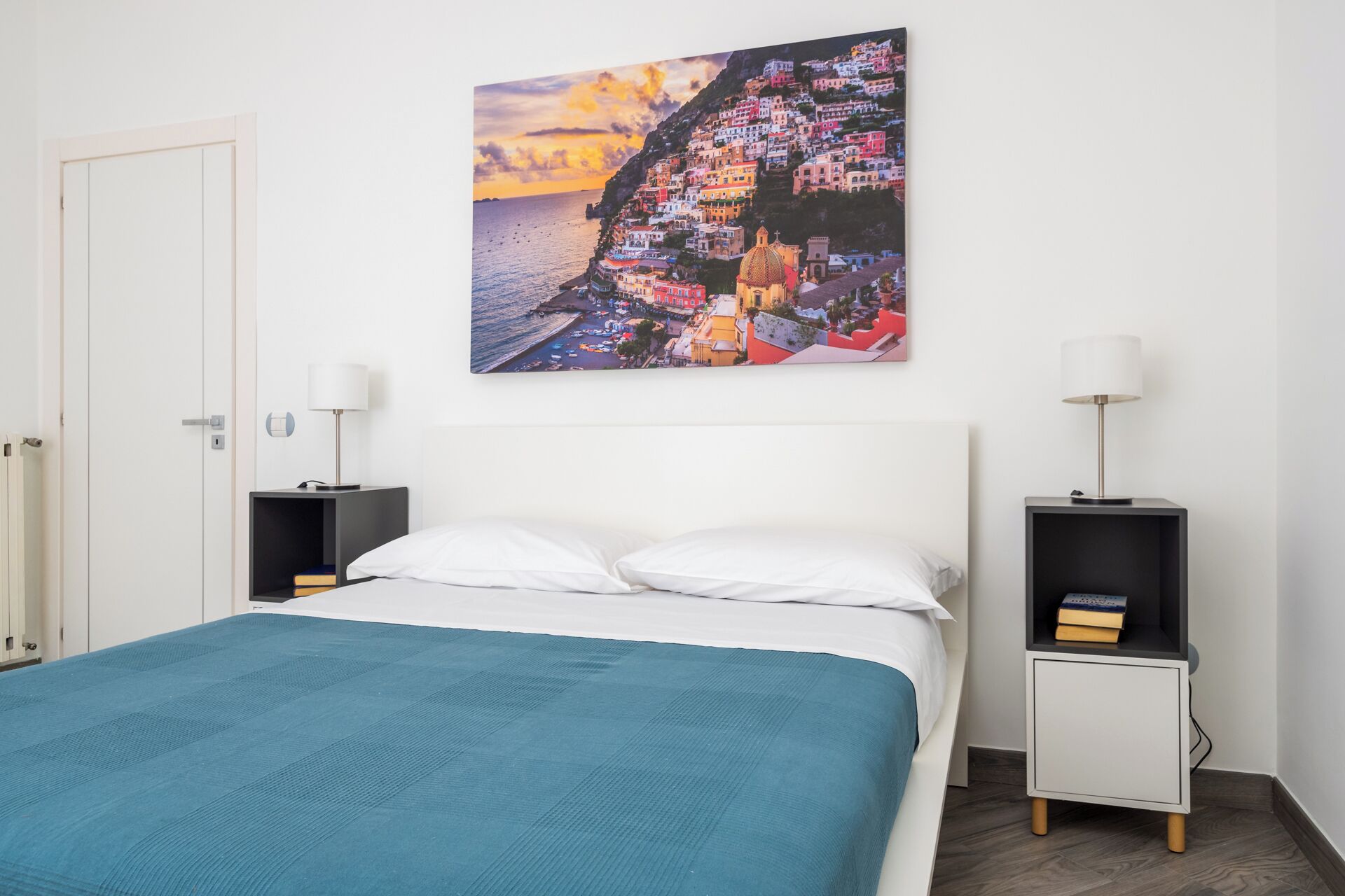 A bedroom with a bed, two bedside tables, a nightstand, and a wall-mounted picture of a coastal town.