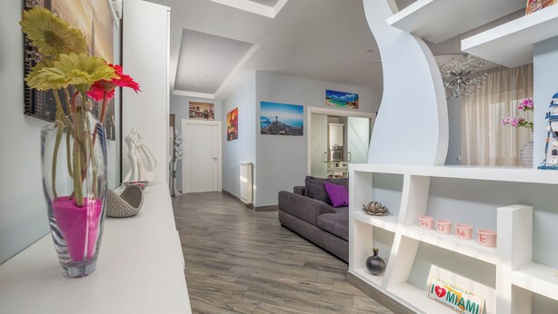 Un salon moderne avec un canapé gris, une console blanche agrémentée d’objets décoratifs et un couloir menant à la cuisine.