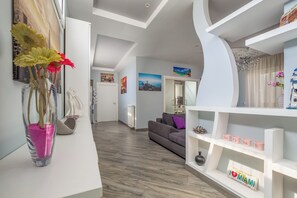 Un salon moderne avec un canapé gris, une console blanche agrémentée d’objets décoratifs et un couloir menant à la cuisine.