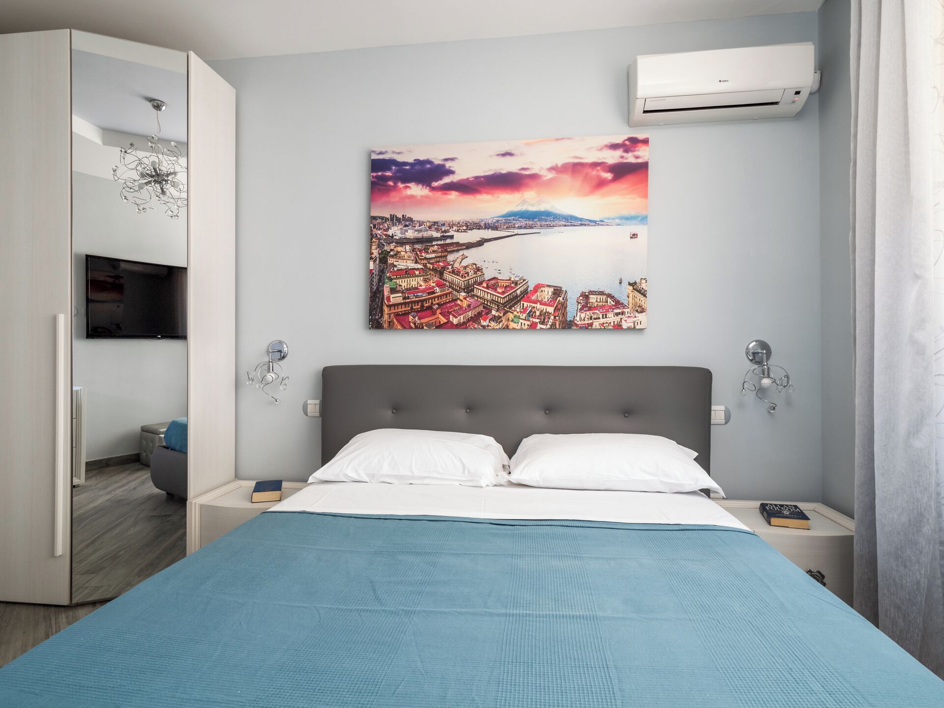 A bedroom with a bed, a wall-mounted TV, an air conditioner, and a cityscape painting.